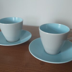 May include: Two light blue and grey ceramic teacups with matching saucers. The cups and saucers are on a wooden surface.