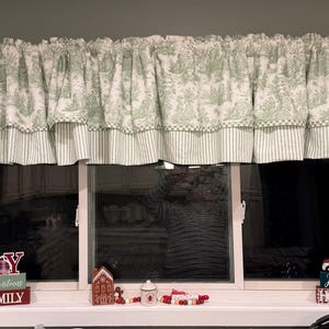 May include: A window valance with a green and white toile pattern, featuring a forest scene. The valance has a ruffled top and a striped bottom border. Decorative holiday signs are on the windowsill below.