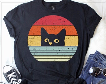 May include: Black t-shirt with a retro sunset design featuring a black cat peeking out from behind the sun.