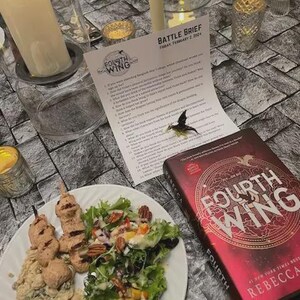 K&ouml;nnte beinhalten: Eine Tischdekoration mit einem Teller mit Essen, einem Buch mit dem Titel "Fourth Wing", Kerzen und einem bedruckten Blatt. Das Buch hat einen roten Einband mit goldenen Buchstaben. Das Blatt tr&auml;gt den Titel "Battle Brief". Der Teller enth&auml;lt Salat und Spie&szlig;e.