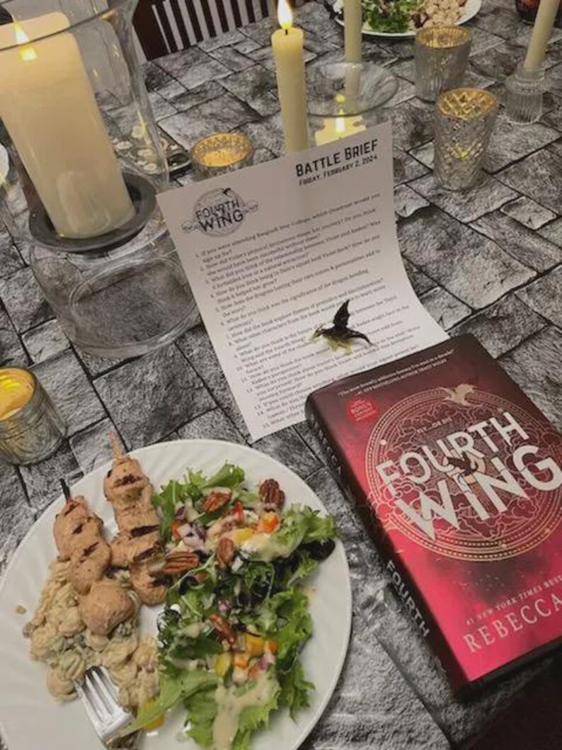 K&ouml;nnte beinhalten: Eine Tischdekoration mit einem Teller mit Essen, einem Buch mit dem Titel "Fourth Wing", Kerzen und einem bedruckten Blatt. Das Buch hat einen roten Einband mit goldenen Buchstaben. Das Blatt tr&auml;gt den Titel "Battle Brief". Der Teller enth&auml;lt Salat und Spie&szlig;e.