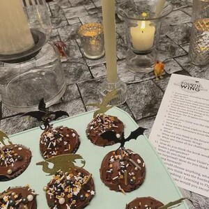 K&ouml;nnte beinhalten: Eine Halloween-Dessert-Pr&auml;sentation mit schokoladen&uuml;berzogenen Leckereien, die mit Streuseln und fledermausf&ouml;rmigen Aufs&auml;tzen verziert sind. Angez&uuml;ndete Kerzen und dekorative Kerzenhalter tragen zur gruseligen Atmosph&auml;re bei.