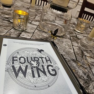 K&ouml;nnte beinhalten: Ein E-Reader mit dem Cover von "Fourth Wing" von Rebecca Yarros, mit einem kreisf&ouml;rmigen Design und dem Text "Fly or Die". Der E-Reader liegt auf einer Oberfl&auml;che mit Steinmuster, umgeben von Kerzen und Kerzenhaltern.