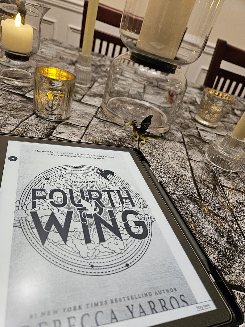 K&ouml;nnte beinhalten: Ein E-Reader mit dem Cover von "Fourth Wing" von Rebecca Yarros, mit einem kreisf&ouml;rmigen Design und dem Text "Fly or Die". Der E-Reader liegt auf einer Oberfl&auml;che mit Steinmuster, umgeben von Kerzen und Kerzenhaltern.