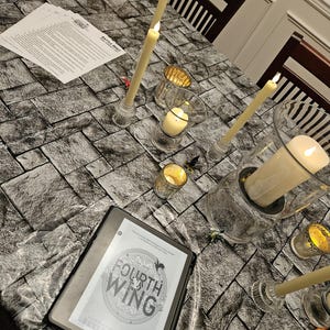 K&ouml;nnte beinhalten: Ein E-Reader mit dem Cover von "Fourth Wing" von Rebecca Yarros, umgeben von brennenden Kerzen in Glashaltern auf einer Tischdecke mit Steinmuster. Die Szene erzeugt eine gem&uuml;tliche, lesefreundliche Atmosph&auml;re.