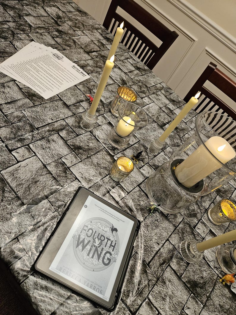 K&ouml;nnte beinhalten: Ein E-Reader mit dem Cover von "Fourth Wing" von Rebecca Yarros, umgeben von brennenden Kerzen in Glashaltern auf einer Tischdecke mit Steinmuster. Die Szene erzeugt eine gem&uuml;tliche, lesefreundliche Atmosph&auml;re.