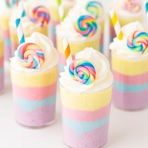 May include: A collection of colorful layered desserts in clear cups, topped with whipped cream and a rainbow swirl lollipop. Each dessert has layers of purple, blue, yellow, and pink. Some have striped straws.