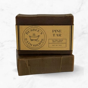 Può includere: Due saponette marroni impilate l'una sull'altra. La saponetta superiore ha un'etichetta di carta marrone su cui è scritto "EST 2018 BUSBEE'S BATH SHOPPE #USETHEFANCYSOAP PINE TAR TEA TREE | RESIN MINT | EARTHY NET WT 5OZ".