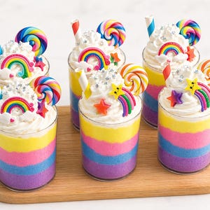 Peut inclure: Six desserts colorés en couches dans des gobelets transparents, disposés sur une planche en bois. Chaque dessert présente des couches aux couleurs de l'arc-en-ciel, de la crème fouettée, un arc-en-ciel, une étoile et une sucette tourbillonnante.