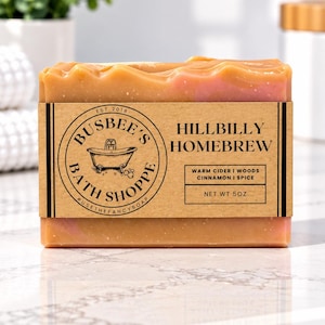 Op de afbeelding: Een rechthoekig stuk oranje zeep verpakt in een bruin papieren label. Het label bevat de tekst Busbee's Bath Shoppe, Hillbilly Homebrew, Warm Cider, Woods, Cinnamon, Spice, Net Wt 5oz en een afbeelding van een badkuip.