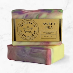 May include: Two bars of handmade soap with a swirling pattern of purple, pink, green, and yellow. The top bar has a brown label that reads "Sweet Pea" and "Delicate & Sweet Floral". The net weight is 5 oz.