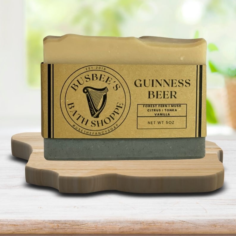 May include: A brown and beige bar of soap with the text 'BUSBEE'S BATH SHOPPE' and 'GUINNESS BEER' on the label. The soap is on a wooden stand. The soap is scented with forest fern, musk, citrus, tonka, and vanilla. The soap is 5oz.