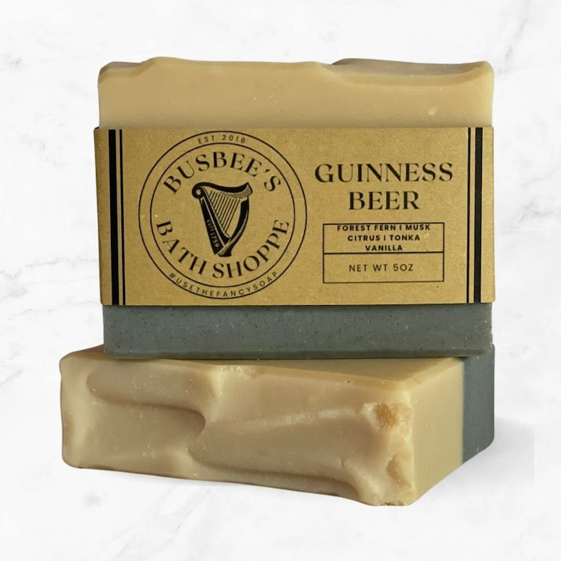 May include: Image of two bars of soap, one on top of the other. The top bar has a brown paper label with black text that reads 'EST 2018 BUSBEE'S BATH SHOPPE #USETHEFANCYSOAP GUINNESS BEER FOREST FERN I MUSK CITRUS I TONKA VANILLA NET WT 5OZ'.