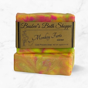 May include: Two bars of handmade soap with a yellow, pink, and green swirl pattern. The soap is labeled "Busbee's Bath Shoppe" and "Monkey Farts scented". The label also states "Cold Process Soap NET WT. approx 4.5 OZ".