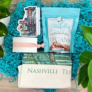 May include: A gift box filled with teal blue crinkle paper. The box contains a tote bag with the text "Nashville Tennessee", a sticker with a drawing of a building with the text "Crossroads Music City", a pink hair tie, and a bag of Southern Praline Pecans from Lottie Belle's.
