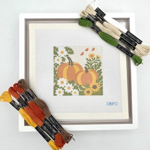 May include: A needlepoint canvas featuring two orange pumpkins surrounded by white and yellow flowers and green foliage. The canvas is framed in a white square frame and accompanied by bundles of embroidery floss in yellow, orange, brown, green, and cream.