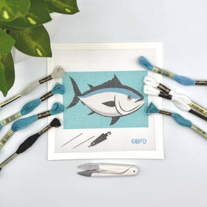 May include: A needlepoint canvas featuring a tuna fish graphic on a light blue background, accompanied by embroidery floss in shades of blue, white, and black, two needles, a needle threader, and white thread snips. The text BPD is printed.