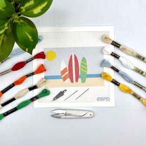 Könnte beinhalten: Eine Stickvorlage mit einer Strandszene, drei Surfbrettern, einer gelben Sonne und einem blauen Ozean. Das Set enthält Stickgarn in Rot, Orange, Grün, Gelb, Weiß und Blau, zwei Nadeln, einen Nadeleinfädler und eine weiße Fadenschere.