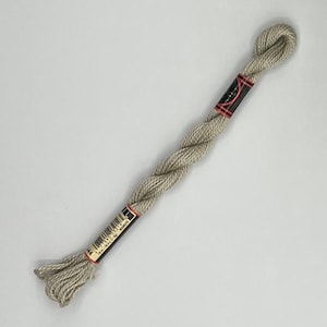 May include: A single skein of light beige embroidery floss, tightly twisted and packaged with black and red labels. The floss is displayed against a plain white background, highlighting its texture and color. Ideal for cross-stitch, embroidery, and other needlecraft projects.