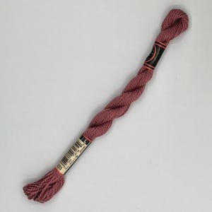 May include: A skein of dusty rose embroidery floss. The floss is tightly twisted and has a label with a barcode and the number 723. The label is black with orange accents. The floss is a muted pink color.