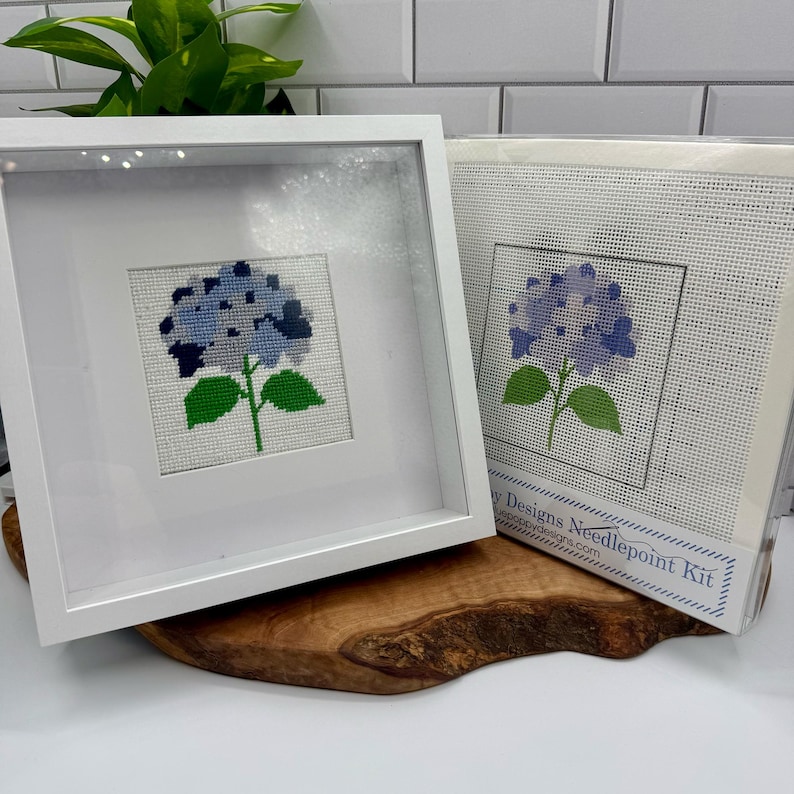 May include: A white square frame containing a cross-stitch of a blue hydrangea flower with green leaves, positioned next to a packaged needlepoint kit featuring the same hydrangea design with the text Poppy Designs Needlepoint Kit.