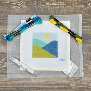 May include: A needlepoint canvas featuring a landscape with a yellow sun, green hills, and blue mountains. Included are two bundles of blue and green embroidery floss, two bundles of yellow and green floss, a needle, and a small mesh square.