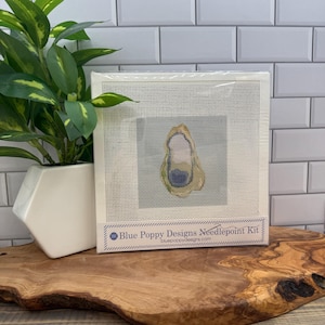 May include: A needlepoint kit with an oyster design. The kit includes a canvas with an oyster image in beige, blue, and yellow. The kit is packaged in a white frame with the text "Blue Poppy Designs Needlepoint Kit".