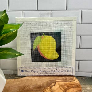 May include: A square needlepoint kit featuring a yellow and green mango graphic on a black background, centered on a white mesh canvas. The bottom label displays the text Blue Poppy Designs Needlepoint Kit and bluepoppydesigns.com.