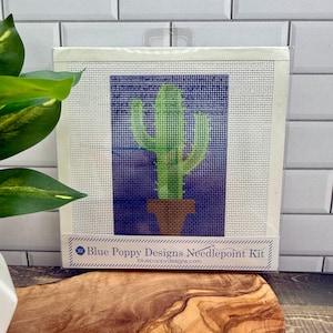 May include: Needlepoint kit featuring a green cactus in a brown pot against a blue background. The kit is packaged in a white frame with the text "Blue Poppy Designs Needlepoint Kit" and the website address.