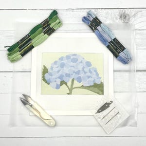 May include: A needlepoint canvas featuring a blue hydrangea flower design, accompanied by two bundles of green embroidery floss, two bundles of blue embroidery floss, a pair of white thread snips, a needle threader, and two sewing needles.