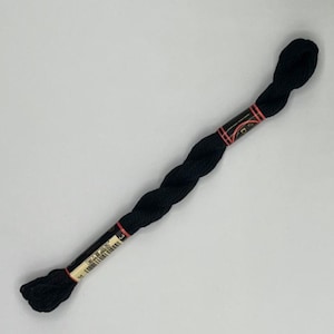 May include: A skein of black embroidery floss, tightly wound and packaged. The floss is a deep, solid black color, with a label wrapped around the center. The label has red accents and text. Ideal for cross-stitch and embroidery projects.
