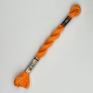 Peut inclure: Une écheveau de fil à broder orange vif, avec une étiquette noire et dorée. Le fil est torsadé et semble prêt à être utilisé pour le point de croix ou d'autres projets de couture. La couleur est un orange vif et uni.