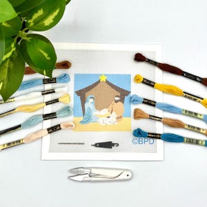 Könnte beinhalten: Eine Stickvorlage mit einer Krippenszene, einem Stall, einem gelben Stern und Figuren, umgeben von verschiedenfarbigen Stickgarnsträngen, einer Nadel und einer kleinen weißen Fadenschere. Auf der Leinwand ist der Text BPD zu sehen.