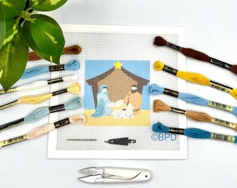 May include: A needlepoint canvas featuring a nativity scene with a stable, a yellow star, and figures, surrounded by various coloured embroidery floss skeins, a needle, and a small white pair of thread snips. The canvas includes the text BPD.