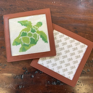 May include: Two square coasters with a stitched turtle design. The turtle is green and white, set against a cream background. The coasters have a brown leather frame. One coaster is face up, the other is face down, revealing the 3M adhesive backing.