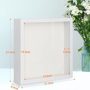 May include: A square white shadow box frame with a clear front panel. The frame has an outer height and width of 21.4 cm, an inner display area of 19.0 cm by 19.0 cm, a depth of 4.4 cm, and an inner depth of 3.0 cm as indicated by measurements.
