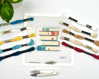 Könnte beinhalten: Eine Stickvorlage mit einem Stapel aus fünf Büchern im Kreuzstichdesign, umgeben von zwölf Stickgarnsträngen in Blau, Gelb, Weiß, Beige und Rot. Ein kleiner Nadeleinfädler aus Metall und eine weiße Fadenschere sind ebenfalls enthalten.