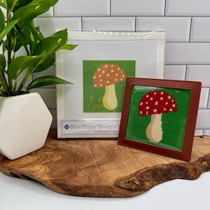 May include: A needlepoint canvas featuring a mushroom design with a light brown cap and white spots on a green background, packaged with a Blue Poppy Designs label, alongside a finished square needlepoint mushroom in a brown leather frame.