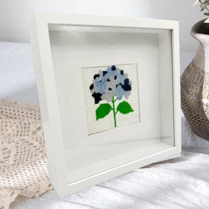 May include: A square white shadow box frame containing a cross-stitch embroidery of a blue hydrangea flower. The flower features shades of light and dark blue petals with a green stem and two green leaves on a white fabric background.