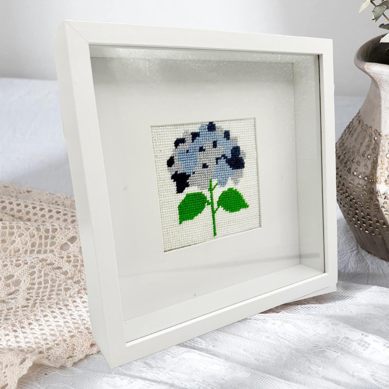 May include: A square white shadow box frame containing a cross-stitch embroidery of a blue hydrangea flower. The flower features shades of light and dark blue petals with a green stem and two green leaves on a white fabric background.