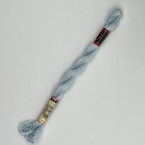 May include: A skein of light blue embroidery floss. The floss is twisted and has a label with the number 775. The label is black and gold with red accents. The floss is a soft, muted blue color.