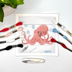 May include: A needlepoint canvas featuring a pink octopus design with light blue bubbles, surrounded by embroidery floss in red, white, light pink, black, and blue. Includes a silver needle, a threader, and white thread snips with the text BPD.