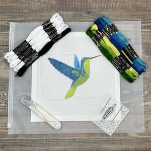 May include: A needlepoint kit featuring a white canvas with a printed hummingbird design in blue and green. The kit includes bundles of white, black, green, yellow, and blue embroidery thread, a white seam ripper, two needles, and a small mesh square.