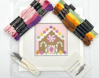 May include: A needlepoint kit featuring a gingerbread house design on canvas, accompanied by two groups of embroidery floss in pink, purple, brown, yellow, green, and white, a pair of white thread snips, and two metal needles.