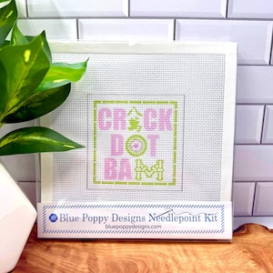 May include: A square needlepoint canvas featuring a grid pattern with the text CRACK DOT BAM in pink and green, including a central green character. The packaging is labeled Blue Poppy Designs Needlepoint Kit with the website bluepoppydesigns.com.