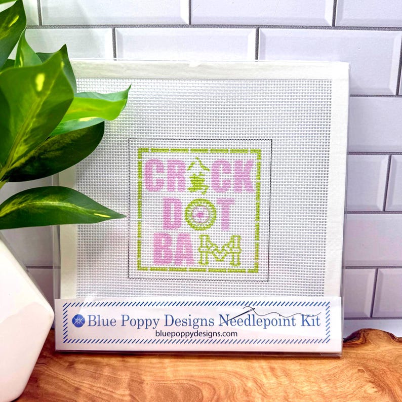 May include: A square needlepoint canvas featuring a grid pattern with the text CRACK DOT BAM in pink and green, including a central green character. The packaging is labeled Blue Poppy Designs Needlepoint Kit with the website bluepoppydesigns.com.