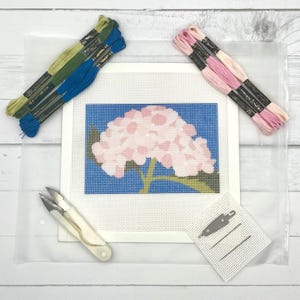 May include: A needlepoint canvas featuring a pink hydrangea flower on a blue background, accompanied by three skeins of green embroidery floss, three skeins of pink embroidery floss, a pair of white scissors, two needles, and a needle threader.