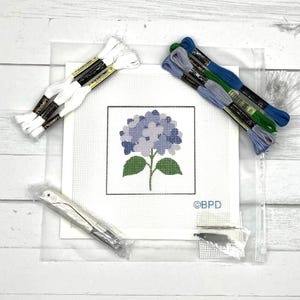Può includere: Un kit per ricamo a mezzo punto con una tela bianca raffigurante un disegno di ortensia stampato in tonalità blu e verdi. Il kit include matassine di filo da ricamo bianco, blu, verde e viola, un ago in metallo e una busta di plastica.