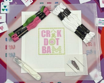 May include: A needlepoint kit featuring a white canvas with a pink and green cross-stitch pattern reading CRAK DOT BAM. Includes green and pink embroidery floss, white embroidery floss, a white seam ripper, two needles, and a small canvas square.