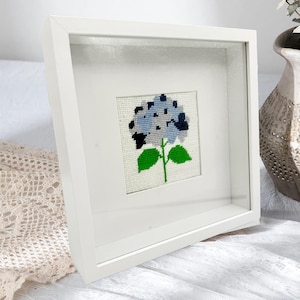 May include: A framed cross-stitch of a blue and black hydrangea flower with green leaves and stem. The artwork is set against a white background and encased in a white, square frame with a glass cover.